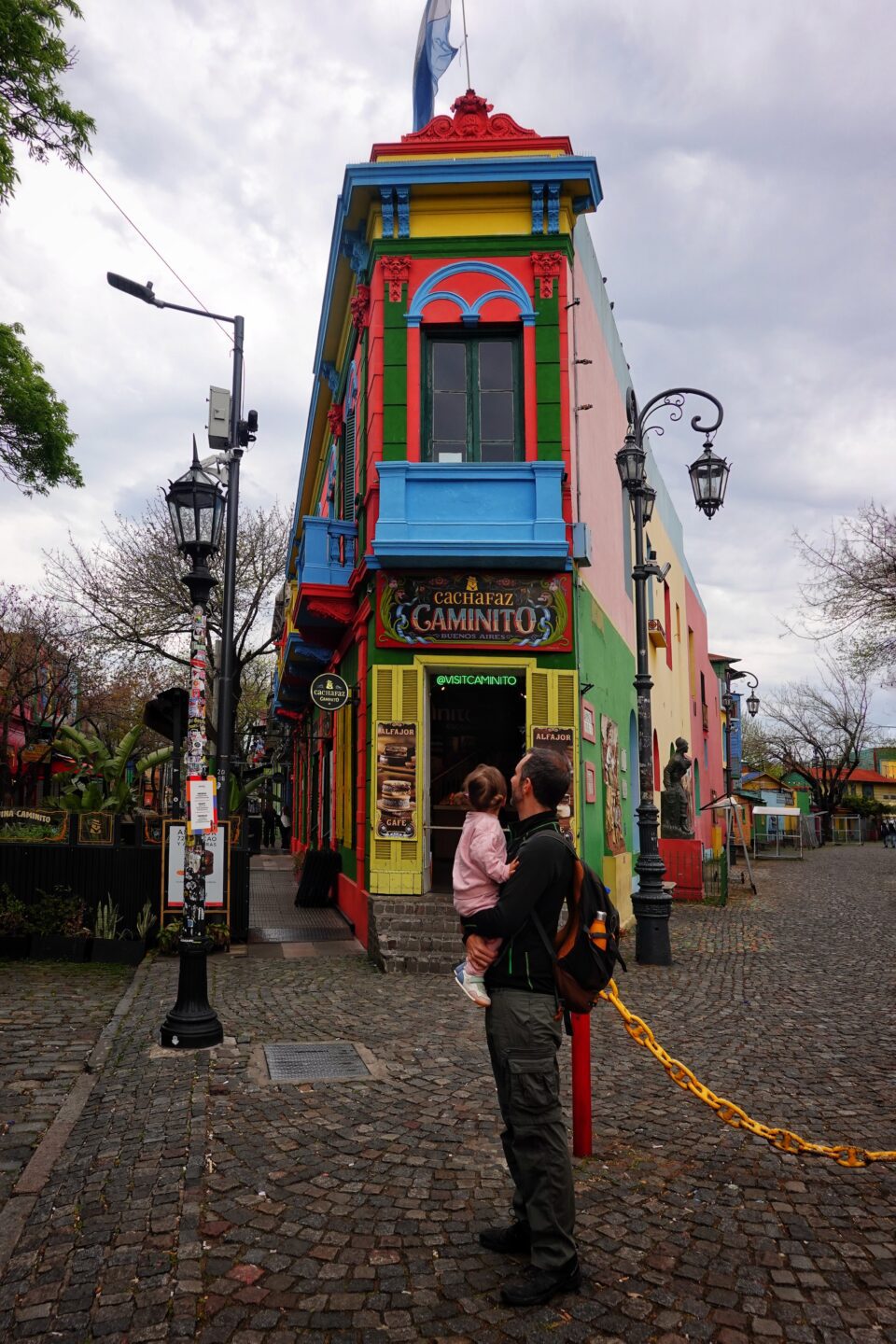 Persona con una bebé en brazos junto a casas de colores en una calle empedrada de La Boca