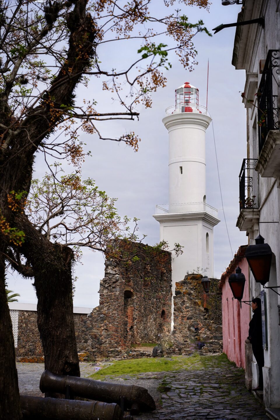 Faro y muralla en ruinas de Colonia del Sacramento vistos desde una calle empedrada, con un cañón antiguo en primer plano