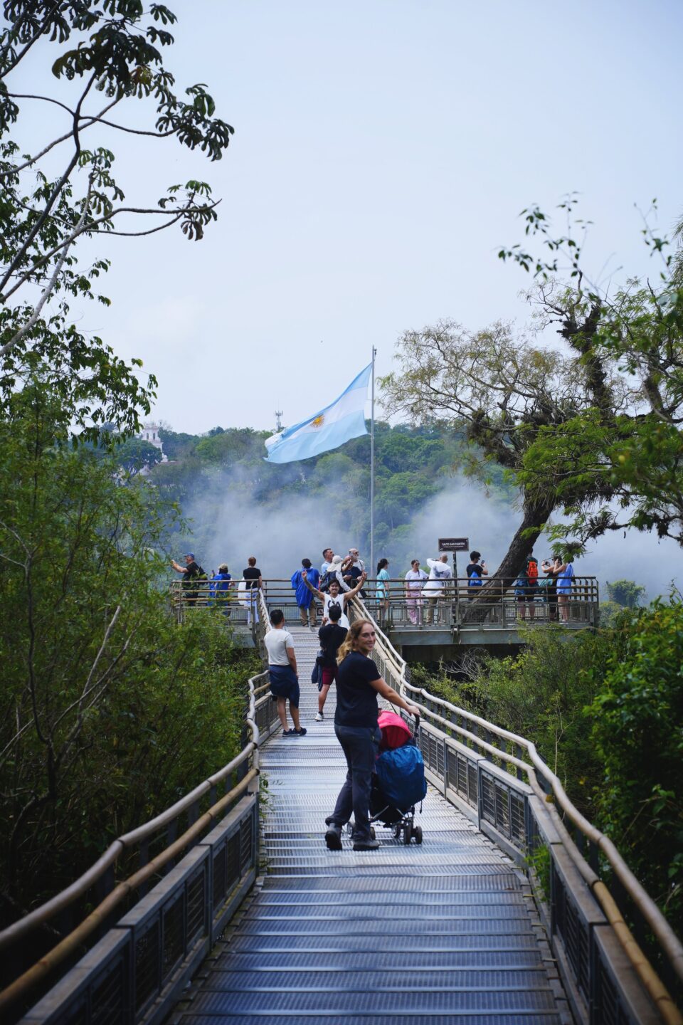Pasarela de madera entre la selva hacia un mirador de las cataratas con gente y una bandera argentina